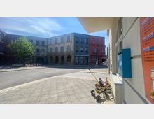 Ein städtischer Platz mit mehrstöckigen Gebäuden im Hintergrund, darunter ein Hotel mit roter Fassade und große Fenster. Im Vordergrund stehen Kinderwagen und kleine Fahrgeräte neben einer Hauswand mit einem Menüplakat.