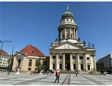 Monumentales Gebäude mit Säulenfassade und großer Kuppel, flankiert von Statuen, vermutlich der Französische Dom in Berlin. Links daneben ein Nebengebäude mit rotem Walmdach, im Vordergrund ein gepflasterter Platz bei klarem, sonnigem Himmel.
