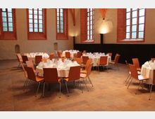 Großer Veranstaltungssaal mit hohen Decken und großen, gotischen Fenstern. Runden Tischen mit weißen Tischdecken und Holzstühlen sind gleichmäßig im Raum verteilt.