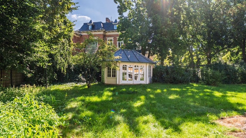 Grüne Gartenanlage mit großer Rasenfläche und hohen Laubbäumen; in der Mitte steht ein heller Pavillon mit vielen Fenstern und achteckigem Dach. Im Hintergrund ist ein mehrstöckiges Backsteingebäude mit Schieferdach teilweise durch das Blattwerk zu sehen.