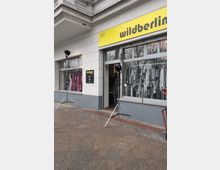 Schaufensterfront eines Modegeschäfts in einem hellen Gebäude mit großem gelben „wildberlin“-Schild über der Eingangstür. Große Fenster zeigen Kleider auf Bügeln, davor stehen zwei Beleuchtungsstative auf dem Gehweg.