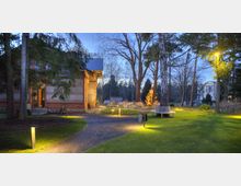 Ein beleuchteter Gartenbereich in der Dämmerung mit einem gepflasterten Weg, Rasenflächen und großen Bäumen. Links befindet sich ein Backsteingebäude, rechts sind Sitzbänke und Tische, und im Hintergrund ist ein weiteres historisches Gebäude sichtbar.