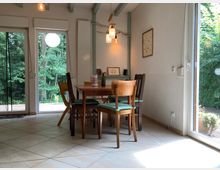 Esszimmer mit Holz-Esstisch und verschiedenartigen Stühlen, umgeben von großen Fenstern, die den Blick auf einen grünen Garten und Wald ermöglichen. Die Wände sind weiß, der Boden gefliest, und die Decke zeigt sichtbare Holzstreben.