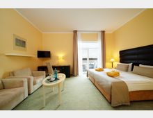 Helles Hotelzimmer mit gelben Wänden, einem großen Doppelbett, zwei Sesseln, einem kleinen Couchtisch sowie einem Schreibtisch mit Stuhl. Auf der Rückseite des Raumes befindet sich eine große Fensterfront mit Zugang zu einem Balkon mit Blick auf das Wasser.