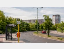 Eine Bushaltestelle mit Wartehäuschen und Sitzbank neben einer leicht kurvigen Straße, umgeben von Bäumen und Grünflächen. Im Hintergrund sind ein mehrstöckiges Wohngebäude und ein moderner Wohnturm zu sehen.