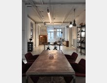 Industriell gestalteter Raum mit hohen Decken, freiliegenden Rohren und weiß gestrichenen Wänden. Im Vordergrund ein großer Tisch mit dunklen Polsterstühlen, im Hintergrund Regale, Holzschränke, und eine Sitzecke vor einem großen Fenster.