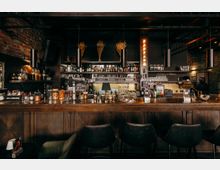 Innenansicht einer Bar im Industrial-Stil mit langer Holztheke, Barhockern und Regalen voller Spirituosen- und Glasflaschen. Sichtmauerwerk, dunkle Decke mit hängenden zylindrischen Lampen und warmes Akzentlicht prägen den Raum.