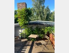 Holzterrasse mit einem kleinen Tisch und drei Stühlen aus Holz und Metall. Im Hintergrund sind grüne Pflanzen, ein Ziegelschornstein und hohe Bäume unter einem blauen Himmel zu sehen.