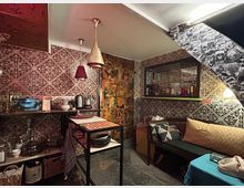 Ein kleiner Raum mit gemütlichem, retro-inspiriertem Dekor. Die Wände sind mit gemusterten Fliesen und Collagen bedeckt, Beleuchtung in Kupfer- und Rotfarben, eine Sitzbank mit Kissen, und eine kleine Küchenecke mit Regalen und Geschirr.