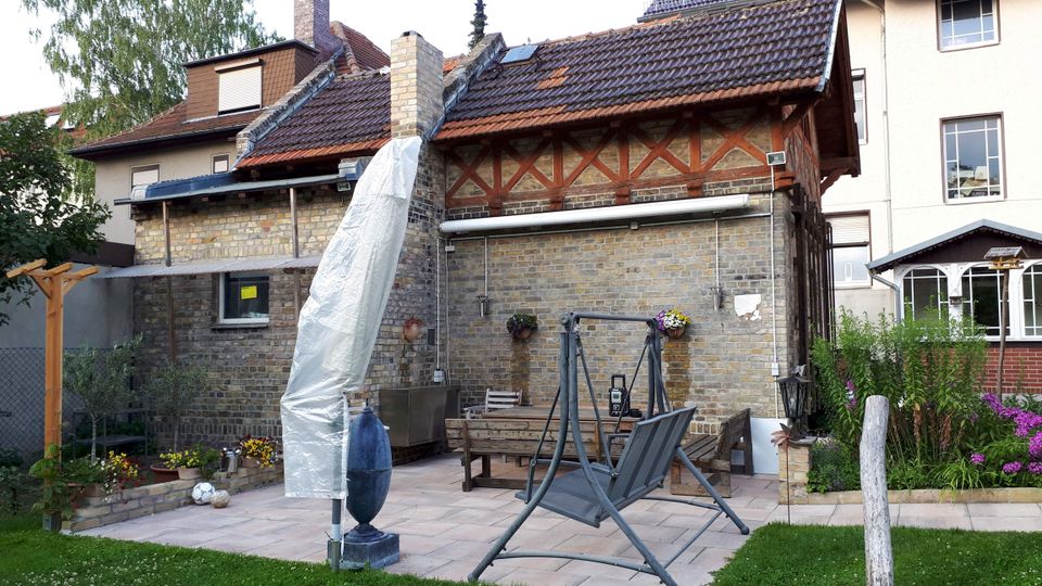 Innenhof mit gepflasterter Terrasse vor einem Backsteingebäude mit Satteldach und sichtbaren Holzstreben unter dem Dach. Auf der Terrasse stehen eine überdachte Gartenliege und Sitzbänke, umgeben von Rasen, Blumenbeeten und angrenzenden Wohnhäusern.