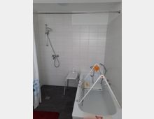 Ein Badezimmer mit weiß gefliesten Wänden und dunklem Boden. Zu sehen sind eine Badewanne mit einem kleinen Wäscheständer darauf, eine Duschstange mit Handbrause an der Wand sowie ein Duschstuhl neben der Badewanne.