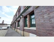 Backsteinfassade eines historischen Industriegebäudes mit großen grün-gerahmten Fenstern. Im Hintergrund ist ein gepflasterter Gehweg, parkende Fahrzeuge und ein Turmgebäude erkennbar.