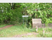 Ein bewaldeter Bereich mit dichtem Grün, einem Schild mit der Aufschrift „Naherholungsgemeinschaft Stiehner See e.V.“, einer Schranke und einem Zaun aus Holz. Im Vordergrund sind Grasflächen und Pfosten mit rot-weißer Markierung zu sehen.