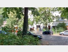 Straßenszene mit Kopfsteinpflaster und parkenden Autos vor einem Wohngebäude mit weißen Fassaden und großen Fenstern. Im Vordergrund sind Bäume und Sträucher, die den Blick auf das Gebäude teilweise verdecken, während ein überdachter Eingang mit Wandgraffiti und ein roter Sonnenschirm sichtbar sind.