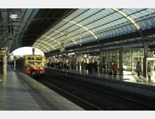 Bahnhof mit überdachten Gleisen und moderner Glasdachkonstruktion, auf einem der Gleise steht eine gelb-rote S-Bahn. Menschen stehen und gehen auf den Bahnsteigen, umgeben von Informationsschildern und gläsernen Wartebereichen.