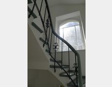 Eine helle Treppenhausansicht mit einer geschwungenen Treppe, Geländern aus schwarzem Metall mit dekorativen Elementen und einem großen, bogenförmigen Fenster im Hintergrund, das Tageslicht einlässt.