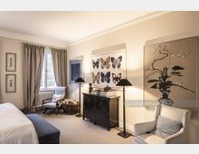 Helles Schlafzimmer mit beigefarbenen Wänden, grauen Vorhängen an einem Fenster, und einem schwarzen Schrank, der von zwei Stehlampen flankiert wird. An den Wänden hängen Kunstwerke, darunter Schmetterlingsbilder und florale Motive, während ein Sessel, ein Korb und ein gepolsterter Sitzhocker das Mobiliar vervollständigen.