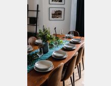 Ein Esszimmer mit einem langen Holztisch, der für mehrere Personen gedeckt ist, einschließlich weißer Teller, Weingläser und einer türkisfarbenen Tischdecke. In der Mitte des Tisches steht ein Blumenarrangement, im Hintergrund sind zwei gerahmte Schwarz-Weiß-Fotografien und eine schwarze Leiter zur Dekoration sichtbar.