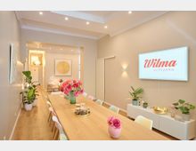 Moderner, heller Veranstaltungsraum mit einem langen Holztisch, umgeben von bunten Stühlen. An der rechten Wand hängt ein Bildschirm mit der Aufschrift „Wilma achtzehn“, umgeben von Pflanzen und dekorativen Elementen; im Hintergrund ein Durchgang mit weiterem Sitzbereich.