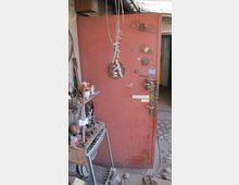 Rote Metalltür in einer Werkstattumgebung, dekoriert mit ungewöhnlichen Objekten wie einer Metallspirale und kleineren Schildern. Daneben stehen Regale mit Werkzeugen, Ersatzteilen und verschiedenen Materialien, während der Boden aus unebenem Kopfsteinpflaster besteht.