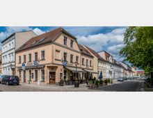 Straßenszene mit gepflasterter Fahrbahn und historischen Gebäuden in Pastellfarben, darunter ein zweistöckiges Haus mit Schildern für „Feinkost“, „Vinladen“ und „Bistro“, sowie Außensitzplätze. Rechts stehen Autos am Straßenrand, links Bäume, die Schatten spenden.