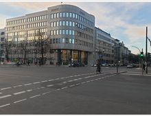 Straßenkreuzung in einer Stadt mit einem modernen Bürogebäude mit abgerundeter Ecke und zahlreichen Fenstern, beschriftet mit "HypoVereinsbank". Im Vordergrund sind Fußgänger, Radfahrer und Autos zu sehen, daneben stehen Straßenlaternen und Verkehrsampeln.