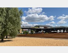 Ein sandiger Platz mit Sitzstufen im Vordergrund, links ein großer Baum. Im Hintergrund eine erhöhte S-Bahn-Brücke mit einem gelben Zug, sowie moderne Gebäude mit Terrassen und Sonnenschirmen. Der Himmel ist blau mit vereinzelten Wolken.