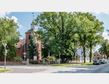 Backsteinkirche mit einem kleinen Glockenturm, umgeben von Bäumen und einem niedrigen Backsteinzaun. Im Vordergrund eine Straßenkreuzung mit Verkehrszeichen, Ampeln und parkenden Autos; im Hintergrund sind Wohnhäuser zu sehen.