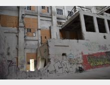 Verlassenes Fabrikgebäude mit beschädigten weißen Wänden, teils zerbrochenen Fenstern und Holzverschalungen. Die Wände sind mit Graffiti bedeckt, und einzelne Metallelemente und offene Bereiche verstärken den ruinösen Zustand.