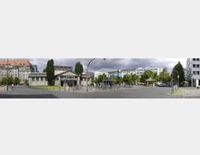 Straßenszene mit einem historischen Gebäude im Zentrum, flankiert von hohen Bäumen. Im Hintergrund stehen mehrstöckige, moderne und historische Wohn- und Geschäftsgebäude. Fahrradständer, Straßenlaternen und Verkehrszeichen sind im Vordergrund sichtbar.