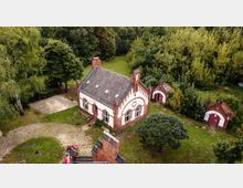 Backsteingebäude im historischen Stil mit weißen Fassaden und roten Fensterrahmen, umgeben von Bäumen und grüner Vegetation. Neben dem Hauptgebäude stehen kleinere, ähnliche Gebäude mit roten Türen, auf einem gepflasterten Hof in ländlicher Umgebung.