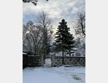 Schneebedeckter Garten mit einer schmiedeeisernen Toranlage in einer gemauerten Steinwand, umgeben von kahlen Bäumen und Nadelbäumen im Hintergrund. Der Himmel ist bewölkt, mit einem Hauch von blauem Himmel sichtbar.