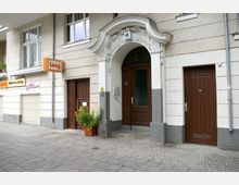 Fassadenansicht eines Altbaus mit verzierten weißen Türrahmen und Hausnummer 45 über der Eingangstür. Links befindet sich ein Café-Schild mit der Aufschrift „Lony Sweets & Coffee“, daneben geschlossene Rolltore, eine braune Holznebentür und ein Pflanzkübel.