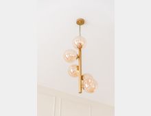 Hängeleuchte mit goldfarbenem Gestell und mehreren kugelförmigen Lampenschirmen aus Glas, montiert an einer weißen Zimmerdecke. Im Hintergrund ist eine Wand mit sichtbaren Vertäfelungen erkennbar.