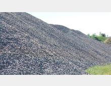 Ein großer Schotterhaufen auf freiem Gelände, rechts im Bild ist eine grüne Wiese mit vereinzelten Büschen und Bäumen im Hintergrund zu sehen. Der Himmel ist klar und hell.