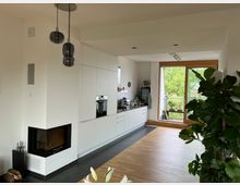 Moderne Wohnküche mit weißen Einbauschränken, integriertem Backofen und Arbeitsbereich. Links befindet sich ein Kamin, während rechts große Fenster mit Holzrahmen und Blick ins Grüne sowie Zimmerpflanzen den Raum prägen.