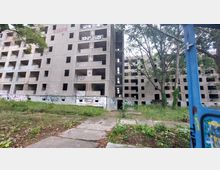 Verlassene, mehrstöckige Betongebäude mit leeren Fensteröffnungen und Graffiti bedeckten Wänden, umgeben von überwuchertem Grün und Bäumen. Im Vordergrund führt ein betonierter Pfad durch die verwilderte Umgebung.