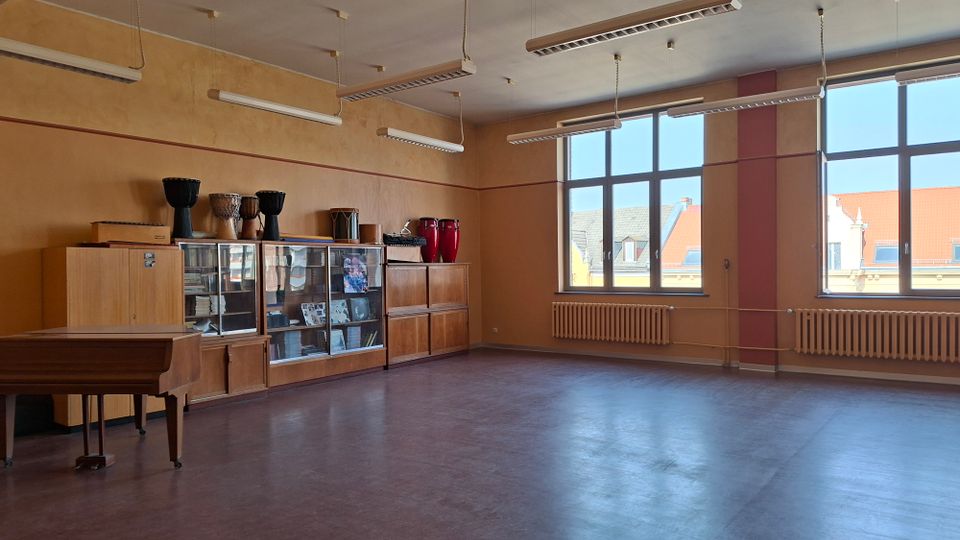Großer, leerer Musik- oder Proberaum mit Linoleumboden, hohen Deckenleuchten und großen Fenstern mit Heizkörpern darunter. An der Wand stehen Holzschränke und Vitrinen mit Instrumenten, daneben ein Klavier; durch die Fenster sind Dächer benachbarter Gebäude zu sehen.