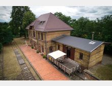 Ehemaliges Bahnhofsgebäude aus gelbem Backstein mit Satteldach, umgeben von Bäumen. Vor dem Gebäude verlaufen alte Bahngleise, und ein gepflasterter Bereich mit Sitzmöbeln und Sonnenschirm bietet eine Terrasse.