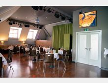 Ein moderner Gastraum mit Holzboden, gedeckten Tischen und braunen Stühlen. Der Raum hat schräge Fenster, eine grüne Trennwand, eine kleine Bühne mit Scheinwerfern und eine Discokugel an der Decke.