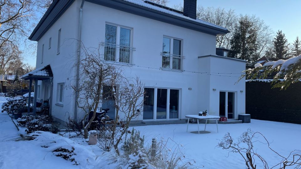 Zweistöckiges, weiß verputztes Wohnhaus mit flachem Walmdach, Schornstein und großen Fensterfronten sowie Schiebetüren zur Terrasse. Im Vordergrund liegt ein verschneiter Garten mit Sträuchern, einem Tisch auf der Terrasse und kahlen Bäumen vor blauem Winterhimmel.