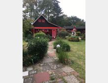 Ein kleines, dekoriertes Haus mit dunklem Holz und roten Akzenten, umgeben von einem gepflegten Garten mit Blumenbeeten, Sträuchern und einem gepflasterten Weg. Im Hintergrund sind hohe Bäume und weitere Grünflächen zu sehen.