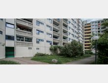 Mehrstöckiges Wohngebäude mit Balkonen und schlichtem, rechteckigem Fassadendesign. Im Vordergrund ein kleiner grüner Innenhof mit Gehwegen, Sträuchern und einer Straßenlaterne.