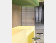 Ein moderner Innenraum mit einer gelben Theke im Vordergrund, zwei chromfarbenen Hockern mit dunklen Sitzflächen und einer Decke mit grünem Lochblech. Im Hintergrund sind eine halbtransparente Wand und bodenlange, schwarze Vorhänge zu sehen.