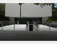 Modernes Gebäude mit einer markanten, weißen, geometrischen Fassade, das auf seitlich gestützten Pfeilern ruht. Im Vordergrund führt eine Rampe mit Geländer zu einem nach unten führenden Eingang, umgeben von gepflastertem Boden und parkenden Autos.