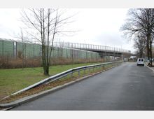 Straßenansicht mit einer schmalen, asphaltierten Fahrbahn und einem Grünstreifen auf der linken Seite, gesäumt von Bäumen und einem Metallgeländer. Im Hintergrund verläuft eine erhöhte Brücke, flankiert von grünen Schallschutzwänden.