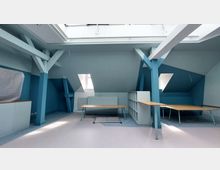 Dachgeschossraum mit hellblauen Wänden und sichtbaren Balkenkonstruktionen, ausgestattet mit simplen Holztischen, Regalen und Dachfenstern, die Tageslicht hereinlassen. Der Boden ist in hellen Blautönen gehalten, und ein Heizkörper befindet sich unter einem der Fenster.