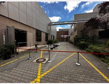 Gepflasterter Innenhof eines Industrie- oder Bürokomplexes mit beige-braunen Gebäudefassaden, rechteckigen Fenstern und sichtbaren Lüftungsrohren oberhalb. Der Bereich ist durch Absperrbänder und Poller abgesteckt, im Hintergrund sind Pflanzen und eine verglaste Gebäudefront zu sehen.