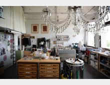 Eine helle Werkstatt mit offenen Regalen, einem großen Holztisch mit Schubladen in der Mitte und diversen Arbeitsmaterialien. Im Vordergrund hängt ein Kristalllüster, und durch die Fenster auf der rechten Seite fällt Tageslicht in den Raum.