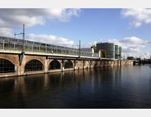Brücke mit Backsteinbögen und darüber verlaufender Bahntrasse neben einem Fluss, im Hintergrund ein modernes Bürogebäude mit Glasfassade und weitere Gebäudestrukturen unter blauem Himmel.